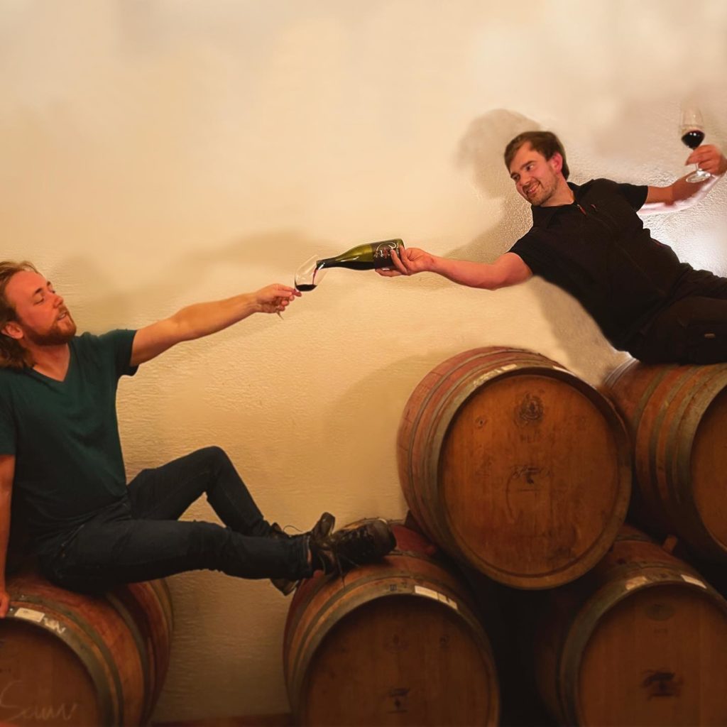 Two happy-looking men, one is Daniel the owner of Hummel Estate, sitting on wine barrels. One is reaching over to wine into the other man´s glass. while holding a wine glass in his other hand.