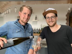 Siegloch Estate Winery. Brothers David and Markus in a vineyard next to each other looking into the camera holding a bottle of wine each.